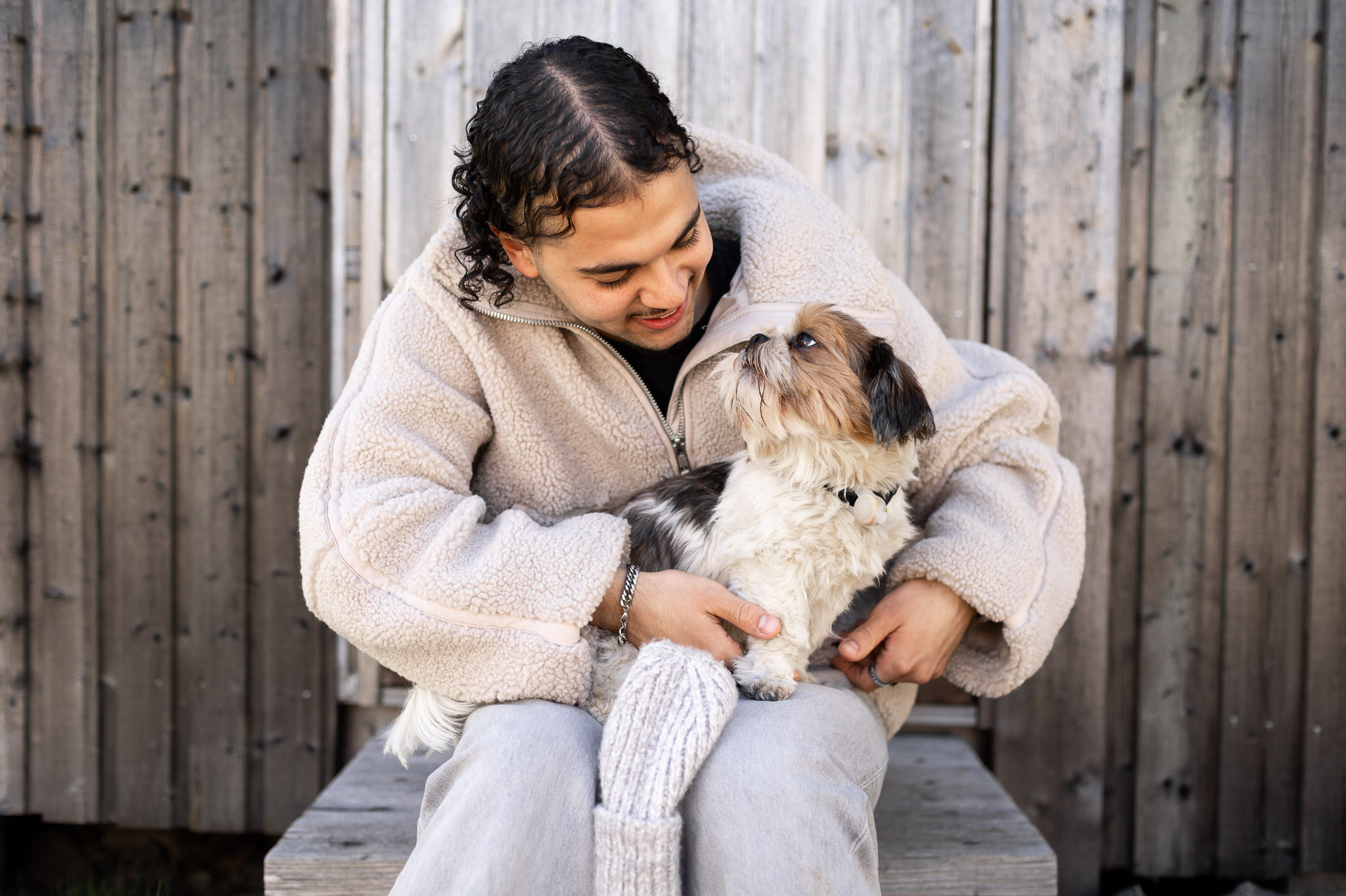 “En person sitter utomhus och håller en liten hund i famnen. Personen ler och tittar kärleksfullt på hunden, som tittar tillbaka.”