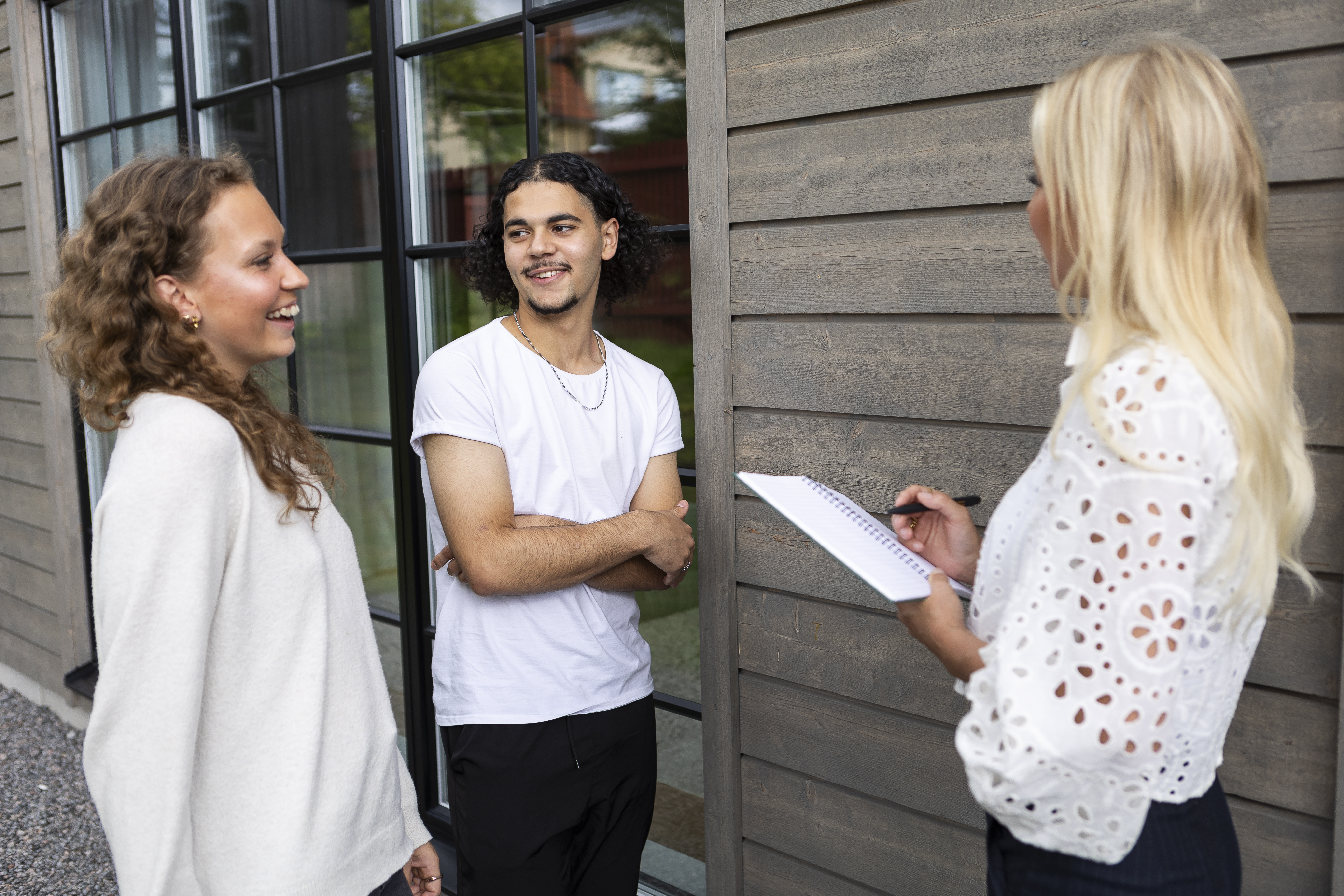 “Tre personer står utomhus och samtalar. Två personer ler och lyssnar medan den tredje håller ett block och penna, som vid en intervju eller ett möte.”