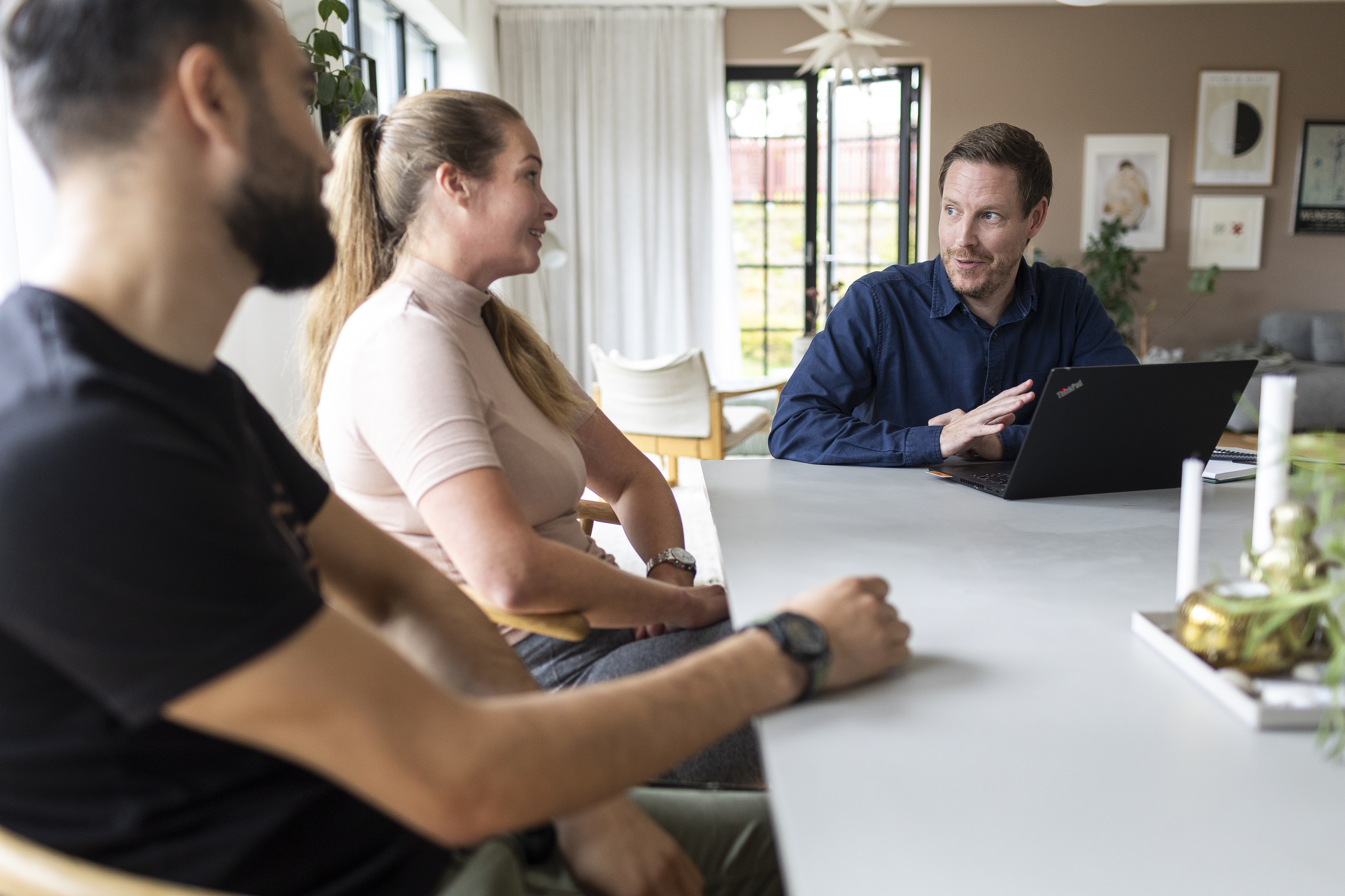 “Tre personer sitter runt ett bord och samtalar. En person har en bärbar dator framför sig och pratar medan de andra två lyssnar och ler.”