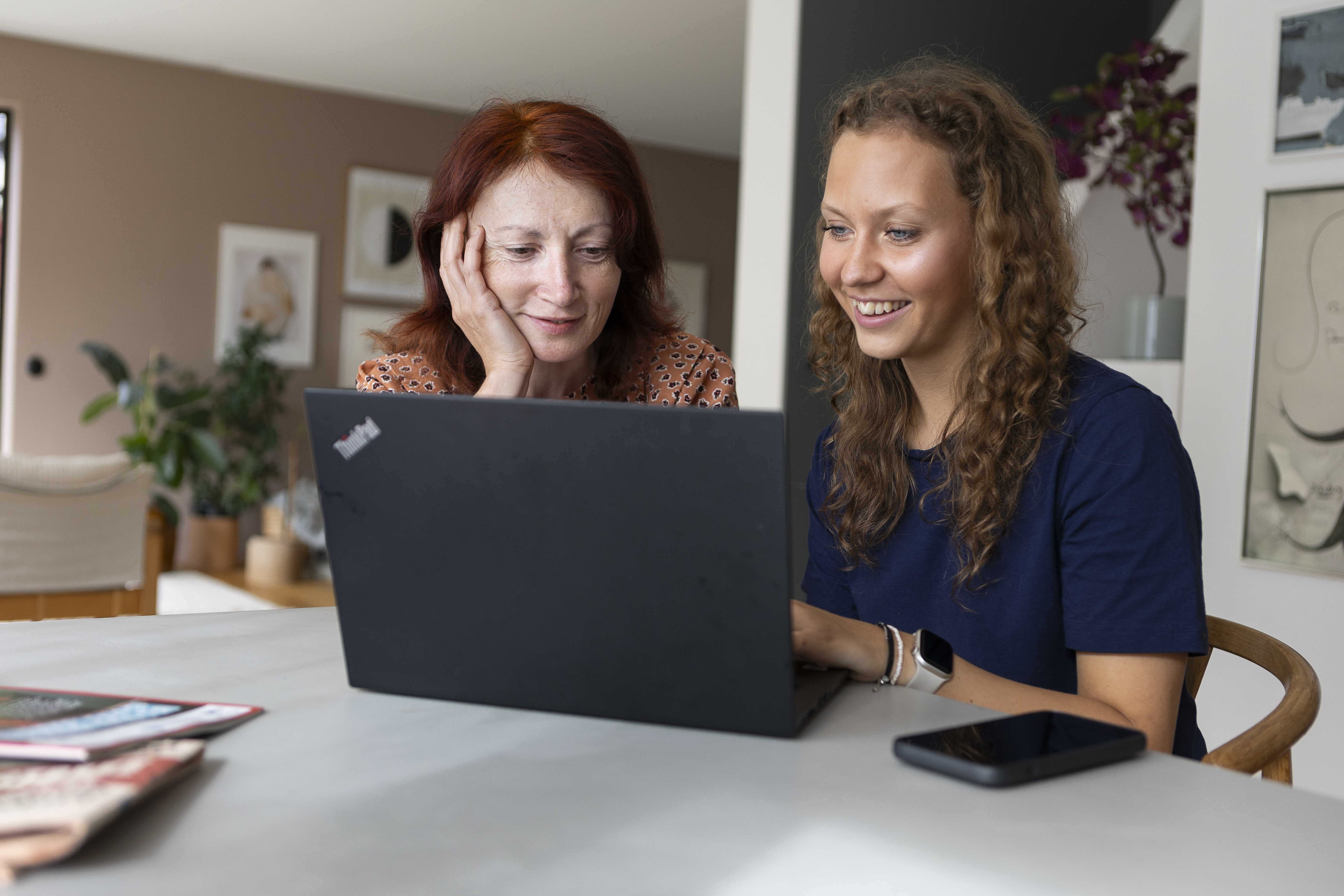 “Två kvinnor sitter vid ett bord och tittar leende på en bärbar dator. Den yngre kvinnan skriver på tangentbordet medan den äldre lutar huvudet mot handen och ser glad ut.”