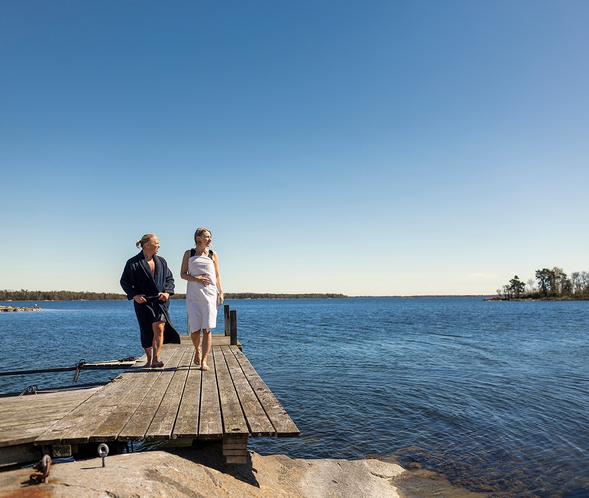 Två personer står på en brygga vid vattnet under klarblå himmel.