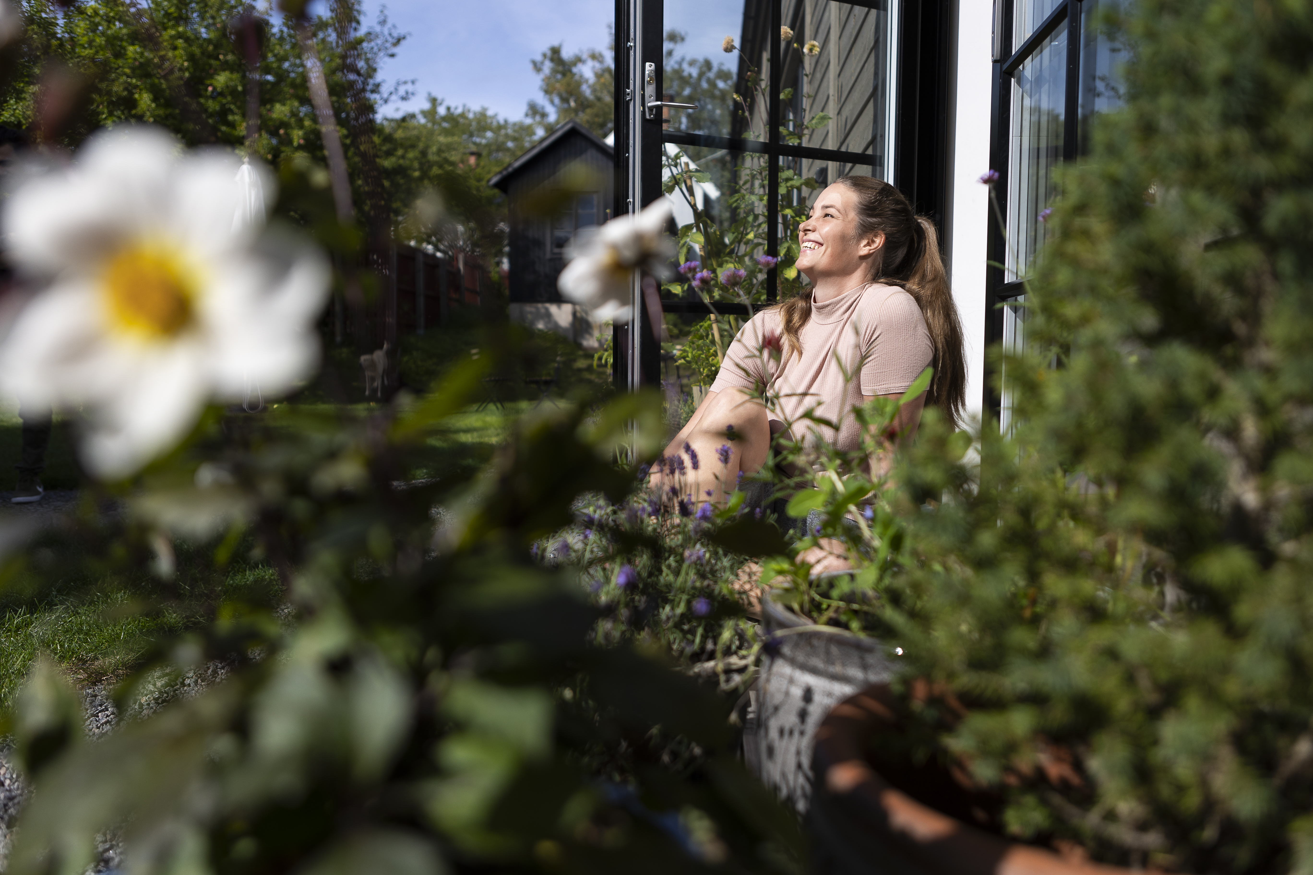 A woman sits outside on a patio surrounded by greenery and flowers, enjoying the sunlight next to a house wall.