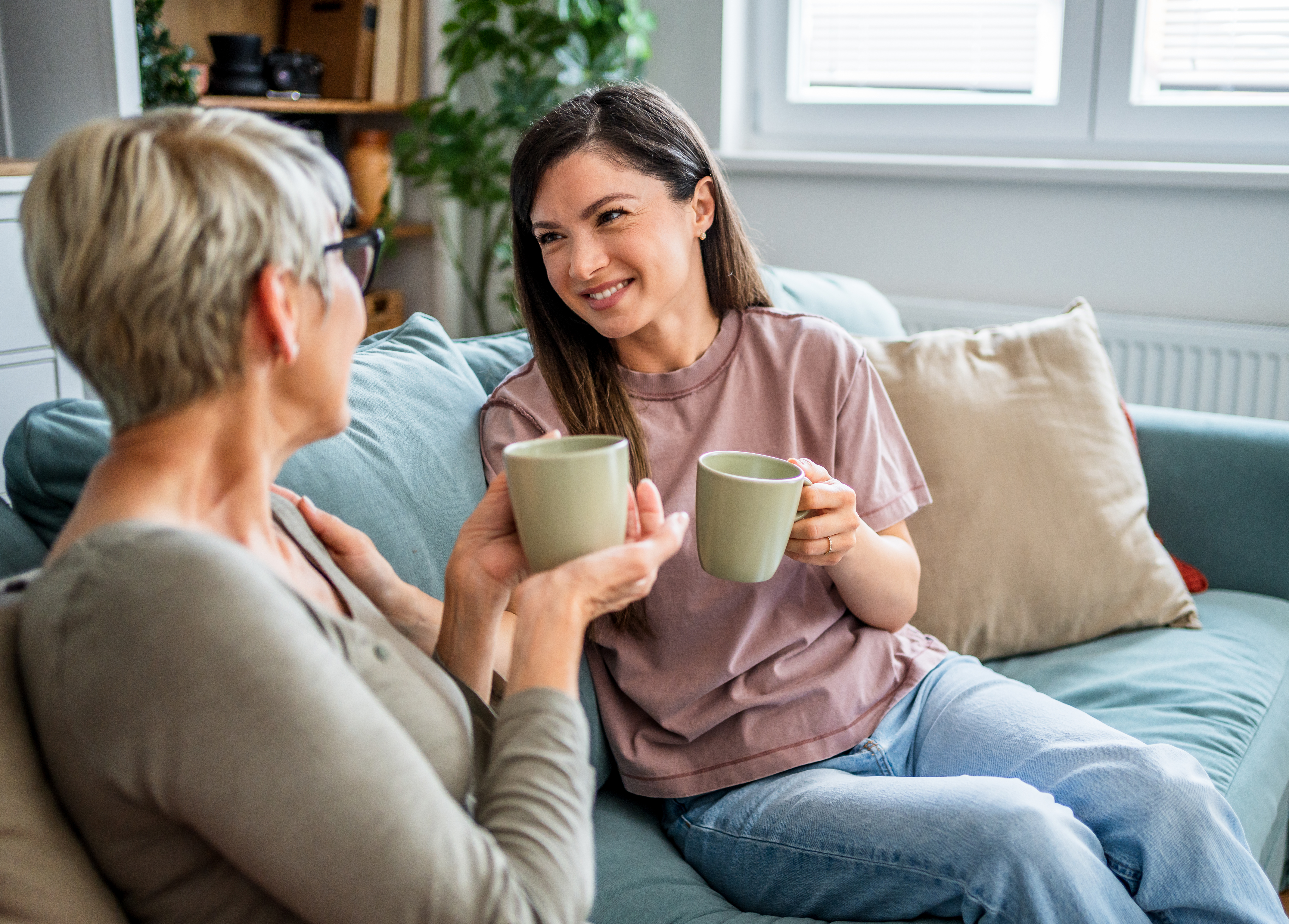 Två kvinnor sitter i en soffa och ler mot varandra medan de håller varsin kopp te eller kaffe, i en varm och avslappnad hemmiljö.