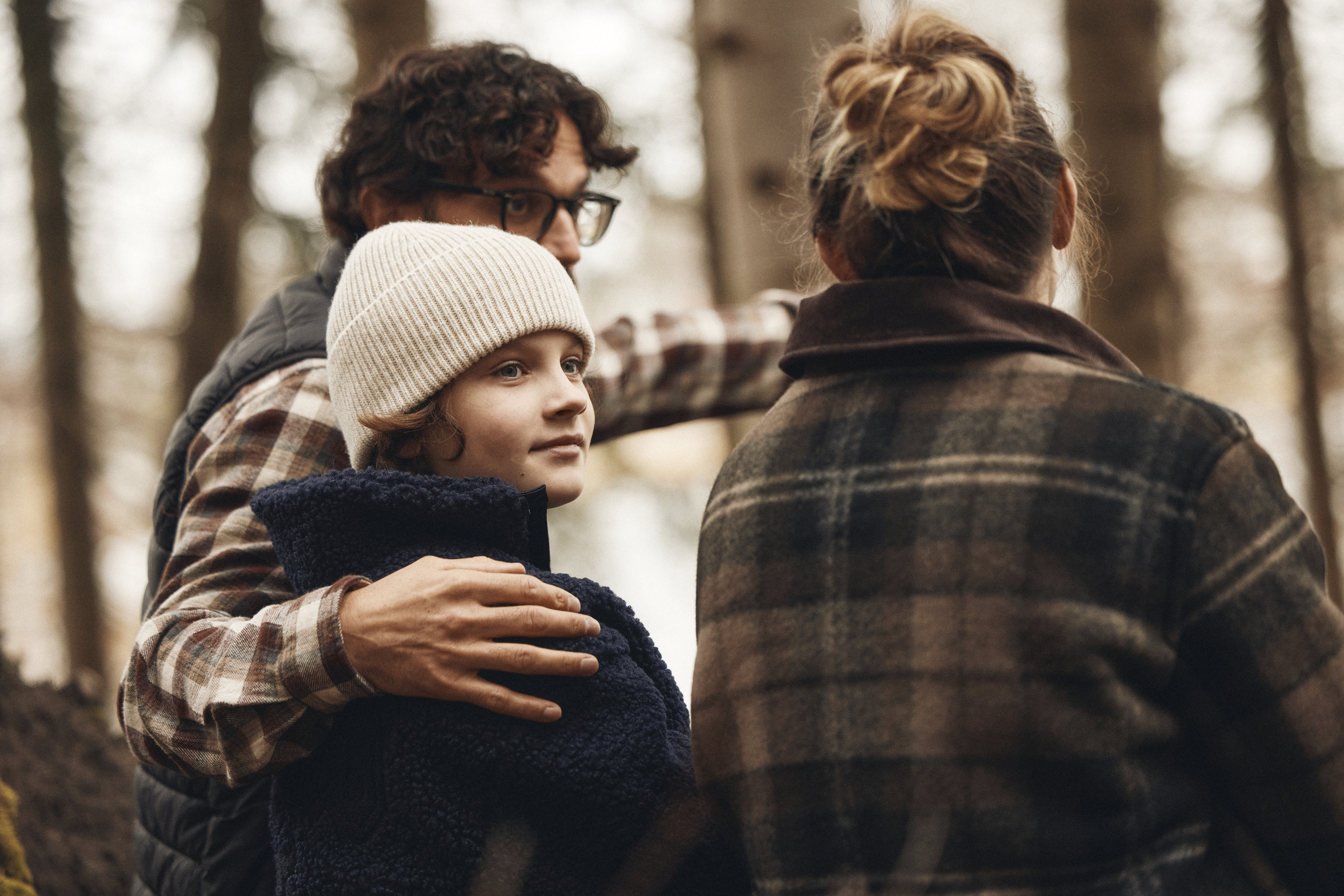 Tre personer står tätt tillsammans i en skogsmiljö. En vuxen håller om ett barn i mössa, medan ytterligare en vuxen står nära – en varm och trygg familjesituation utomhus.