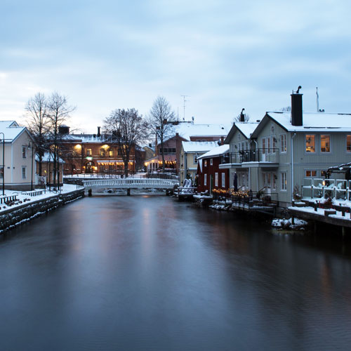 Vintervy över ett stilla vattendrag i stadsmiljö med bro, hus längs kajen och snö på marken i skymningsljus.