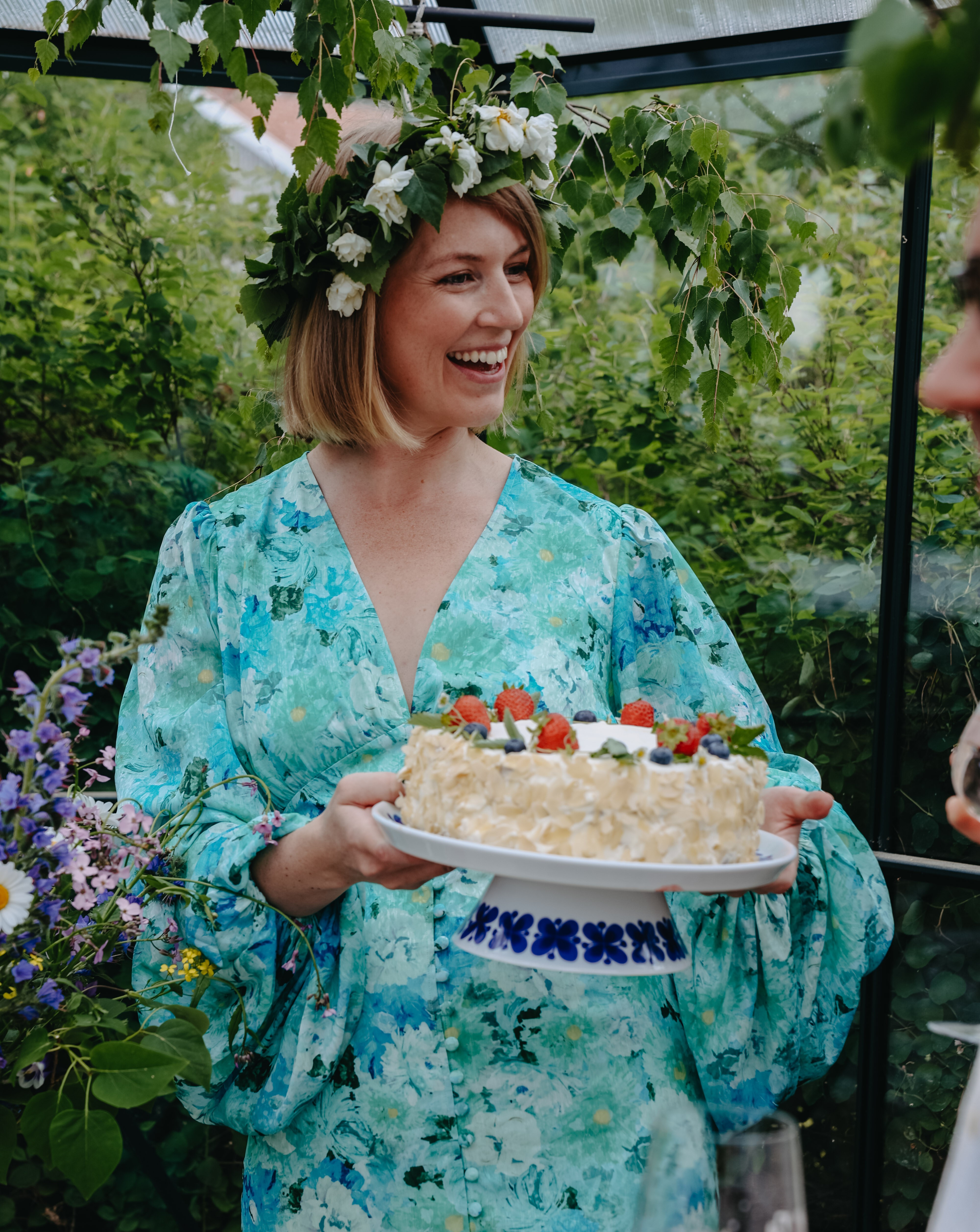 En leende person i ljusblå blommig klänning står i ett grönskande växthus och håller en gräddtårta dekorerad med jordgubbar och blåbär. Personen bär en blomsterkrans med vita blommor och gröna björkkvistar, omgiven av sommarblommor och frodig grönska.
