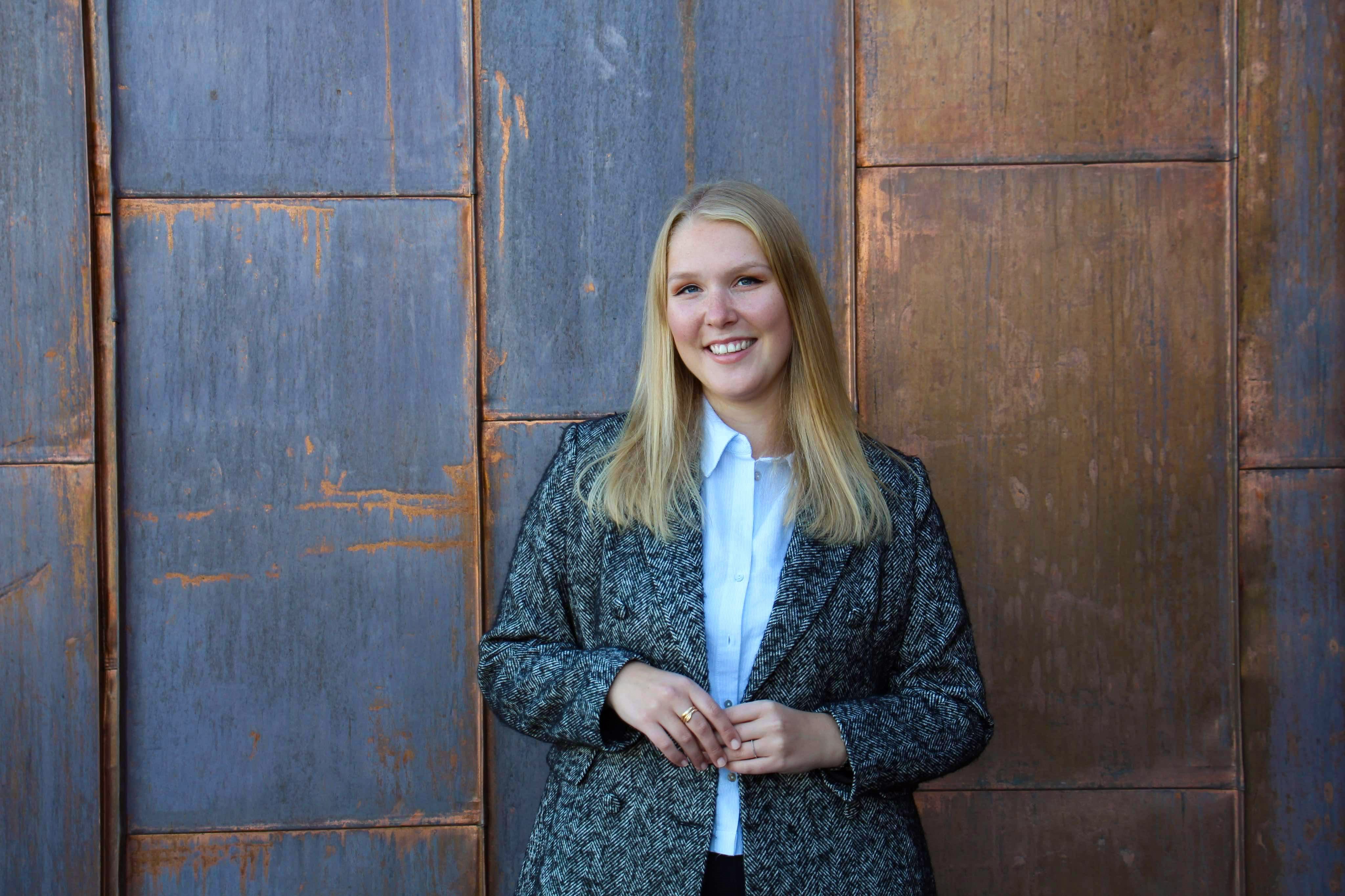 Leende person med långt blont hår står framför en vägg av rostfärgade metallpaneler, iklädd vit skjorta och mörk mönstrad kavaj.