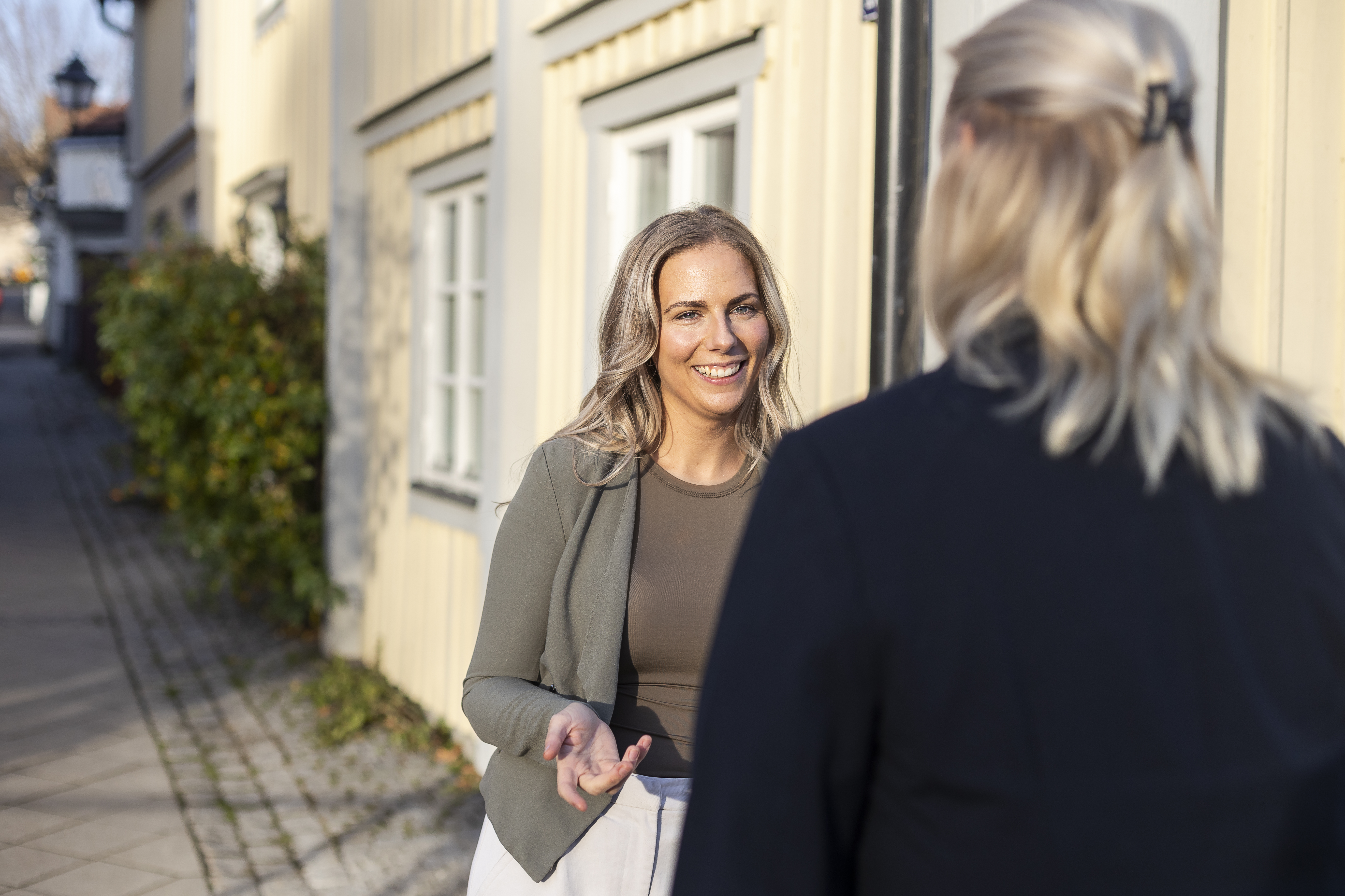 Två personer samtalar utomhus utanför en ljus träbyggnad. En leende bankrådgivare står vänd mot kameran och pratar med en kund som syns bakifrån i kvällssolen.
