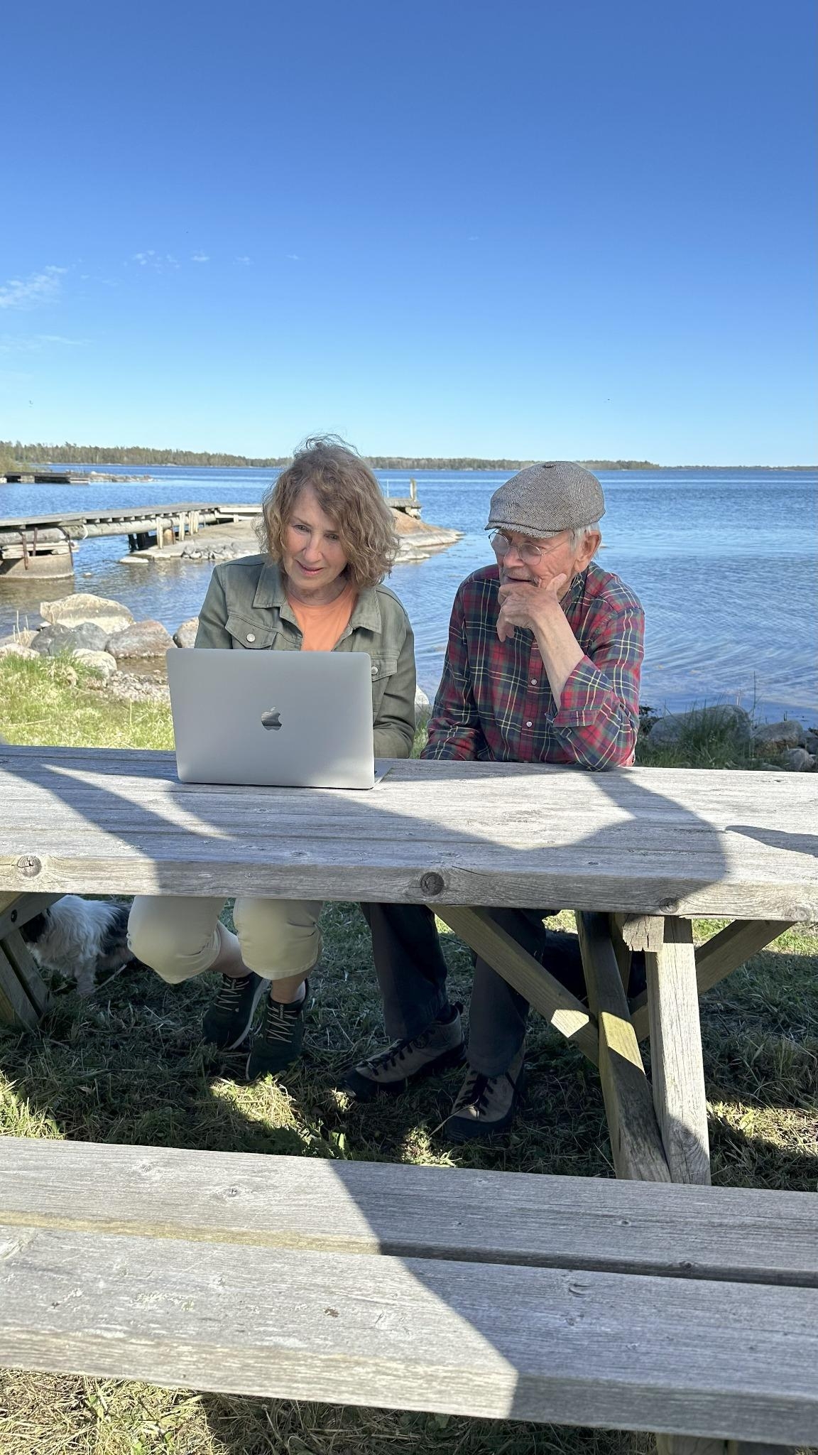 Två äldre personer sitter vid ett träbord utomhus vid vattnet och tittar tillsammans på en bärbar dator. I bakgrunden syns en brygga, klippor och hav med klarblå himmel.