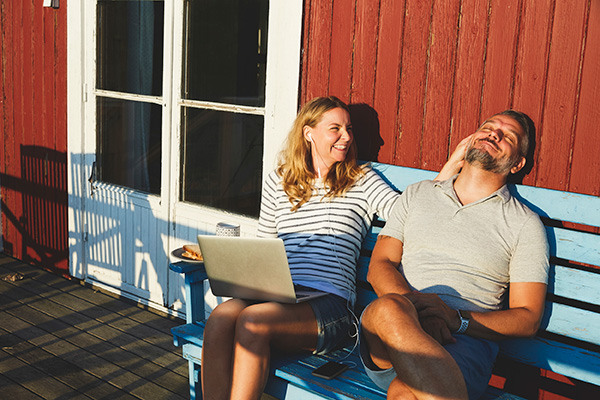 Två personer sitter på en blå bänk i solen utanför ett rött trähus. Den ena personen har en laptop i knät och ler mot den andra, som lutar sig bakåt och skrattar. De verkar njuta av en avslappnad stund tillsammans.