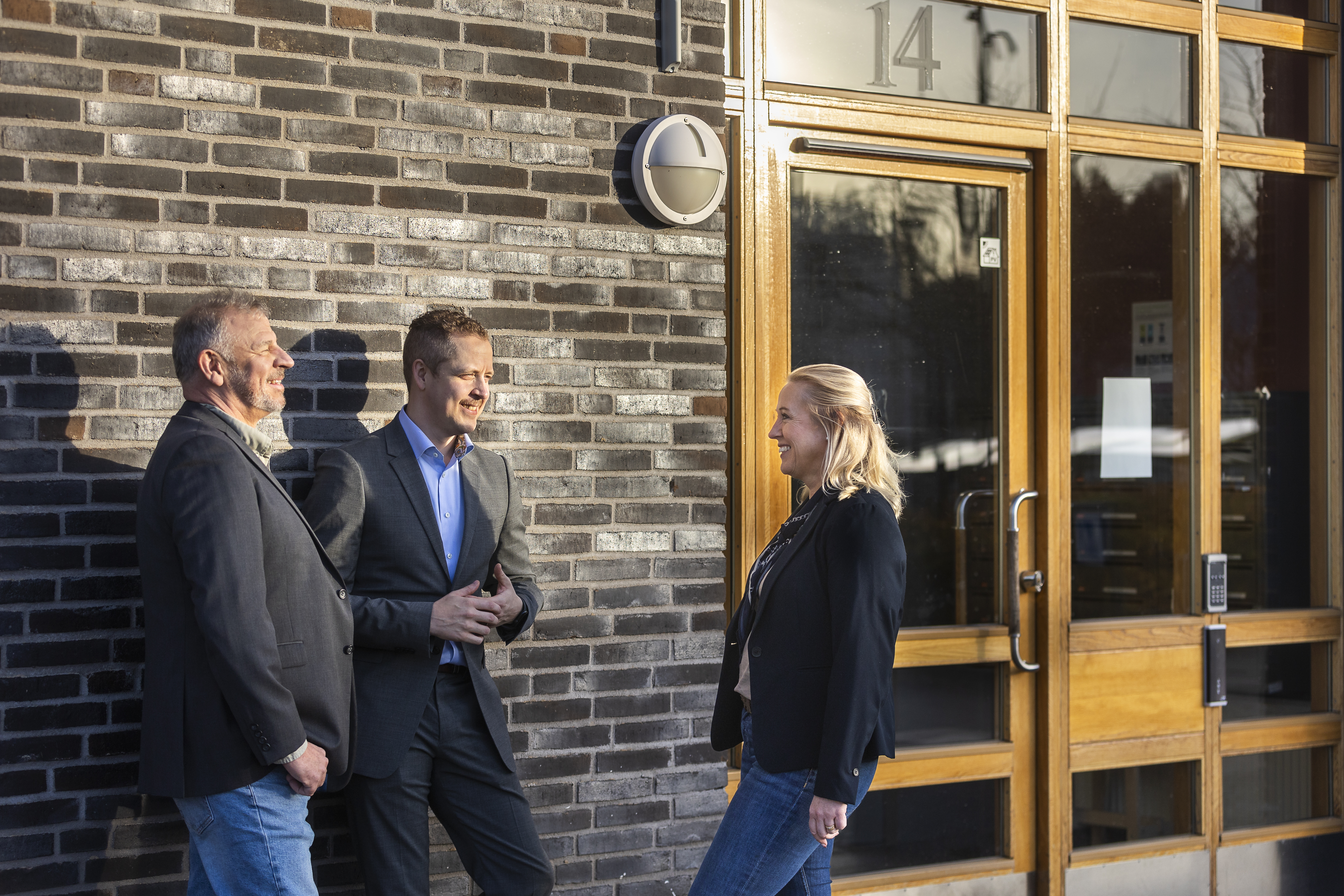 Tre av bankens rådgivare står utanför en byggnad med nummer 14 vid entrén. Två manliga rådgivare i kavaj och en kvinnlig rådgivare i kavaj och jeans pratar och ler mot varandra i solskenet, framför en tegelvägg och en trädörr med glaspartier.